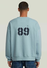 Sudadera de color azul claro con un corte oversized, que presenta un gran '89' impreso en negro en la parte posterior. Textura suave y puños acanalados.
