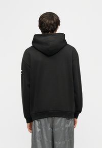 Homme de dos portant un sweat à capuche noir et un pantalon gris ample sur un fond clair uni.