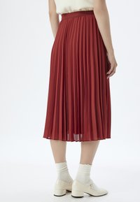 Roter plissierter Rock mit glatter Textur, hoher Taille und knielangem Design, kombiniert mit weißen Socken und glänzenden Blockabsatzschuhen.