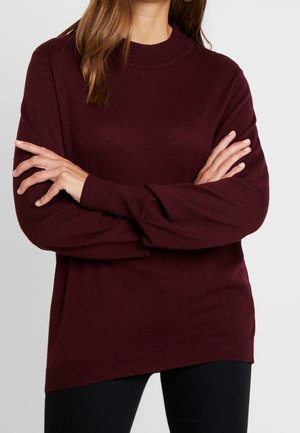Femme portant un pull à manches longues de couleur bordeaux foncé avec un col montant, bras croisés sur la poitrine, devant un fond clair uni.