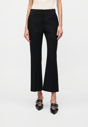 Femme portant un pantalon flare court noir et des mules noires à bout pointu avec des boucles argentées, debout devant un fond uni.