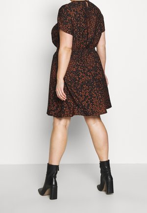 Kvinde iført en knælange sort kjole med orange leopardprint og sorte ankelstøvler, stående mod en ensfarvet hvid baggrund.