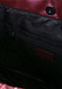 Indvendig i en maroon taske med sort foer. Har en lynlåslomme og et prægget logomærke. Glatte og strukturerede materialer er til stede.