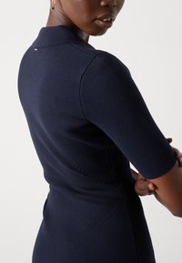 Femme aux bras croisés portant un haut en maille ajusté bleu marine à manches courtes, montrant les détails côté et dos sur un fond uni.