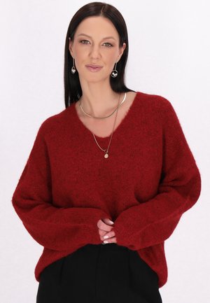 Maglione rosso a maglia con scollo a V, vestibilità ampia e maniche lunghe. Texture morbida con una finitura leggermente pelosa. Indossato su pantaloni neri.
