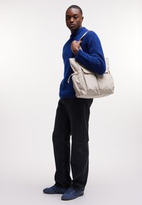 Sac beige en matériau doux et texturé avec un cordon de serrage, porté sur un pull bleu en peluche et associé à un jean noir et des chaussures bleues.