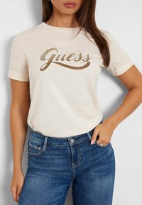 Camiseta de algodón color crema con mangas cortas enrolladas y logo "Guess" brillante en dorado metálico. Combinada con unos jeans de mezclilla azul.
