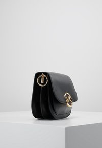 Mustaa nahkaista crossbody-laukku kultaisella pyöreällä soljella ja avaimenperällä, esittäytyy valkoisella pylväällä harmaata taustaa vasten.