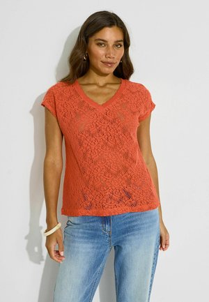 Mujer con una blusa naranja de encaje de manga corta y vaqueros azules, de pie frente a un fondo claro y liso.