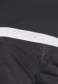 Džíny Calvin Klein s černou denimovou tkaninou, s kontrastním bílým pasem označeným embosovaným textem loga.
