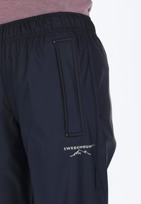 Marineblau Wasserfeste Hose mit elastischem Bund, Seitentasche und einem in Weiß bedruckten Logo. Das Material hat eine glatte, glänzende Textur.