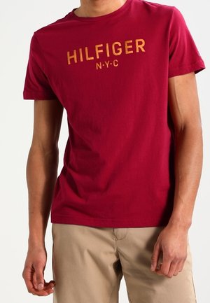 Mężczyzna w czerwonej koszulce z krótkim rękawem z napisem "HILFIGER N·Y·C" w żółtym kolorze i beżowych spodniach, stojący z rozluźnionymi rękami.