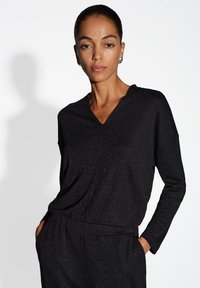 Top nero a V con maniche lunghe, realizzato in tessuto testurizzato con un sottile luccichio. Presenta una vestibilità rilassata e tasche laterali.