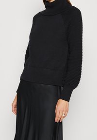 Schwarzer Rollkragenpullover mit strukturierten Ärmeln und gerippten Bündchen, kombiniert mit einem glatten schwarzen Satinterock.