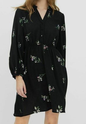 Czarna sukienka z długim rękawem z małymi kwiatowymi wzorami, zapinana na guziki z przodu, zebrana w talii i z kołnierzykiem, noszona przez stojącą osobę.