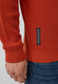 Strukturierter orangefarbener Pullover mit gerippten Bündchen und Saum. Auf der Seite befindet sich ein Etikett mit der Aufschrift "FÜR ALLE BEDINGUNGEN GEMACHT". Getragen mit blauen Jeans.