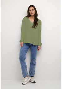 Lichtgroene blouse met lange mouwen en een V-hals, gecombineerd met blauwe spijkerbroeken en witte sportschoenen, staand tegen een neutrale achtergrond.