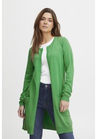 Cardigan vert en tricot doux, avec une coupe longue, un devant ouvert, des manches longues et deux poches avant. Porté par-dessus un haut blanc.