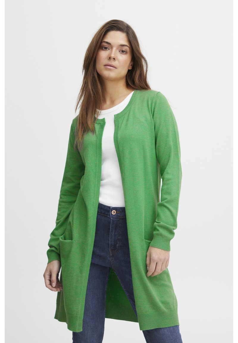 Cardigan vert en tricot doux, avec une coupe longue, un devant ouvert, des manches longues et deux poches avant. Porté par-dessus un haut blanc.