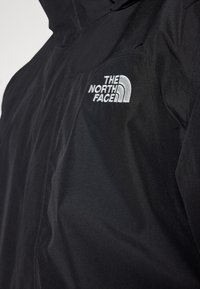 Černá nepromokavá bunda s vysokým límcem, zipovým zapínáním a vyšívaným bílým logem The North Face na levé části hrudi.