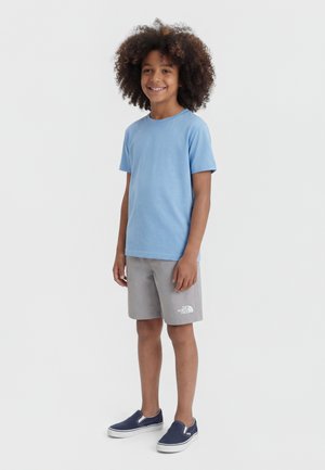 Naeratav laps lokkis juustega, kandes helesinist t-särki, halli The North Face lühikest pükst ja mereväesiniseid libisevaid kingi, seisab valgel taustal.