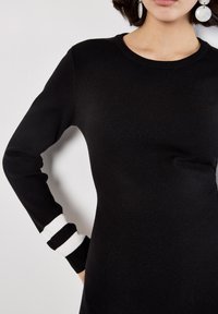 Schwarzes langärmliges Kleid mit taillierter Passform. Verfügt über weiße Streifen an den Bündchen. Hergestellt aus einem glatten, gerippten Strickmaterial.