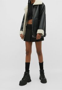 Chaqueta de cuero negra con cuello de borrego sintético blanco, combinada con una falda negra con pliegues y botas negras de tobillo robustas. Medias hasta la rodilla negras.