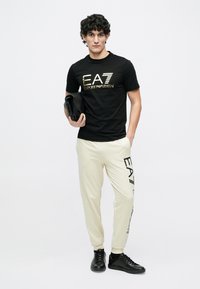 Fiatal férfi fekete EA7 Emporio Armani pólóban és világos színű szabadidőnadrágban, aki egy fekete bőr borítéktáskát tart egyszerű fehér háttér előtt.