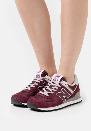 Osoba nosząca bordowo-białe sneakersy New Balance 574 z białymi sznurówkami, stojąca na jednolitym białym tle.