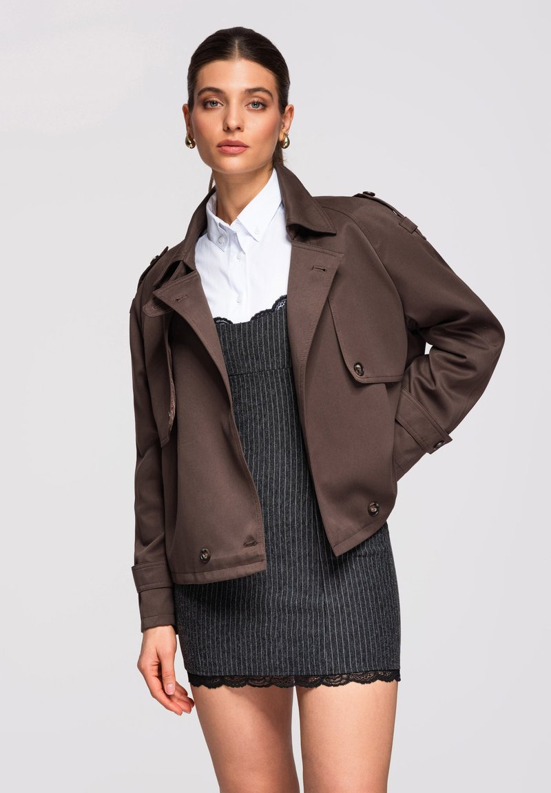 Femme portant un manteau court marron sur une chemise blanche et une robe noire à rayures fines avec un ourlet en dentelle, posant avec la main sur la hanche devant un fond uni.