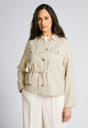 Frau mit langen dunklen Haaren, die eine beige, geknöpfte Jacke mit Vorderentaschen und Taillenband trägt, kombiniert mit weißen Hosen.