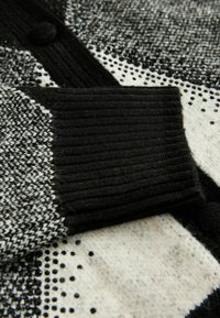 Černobílý pletený cardigan s žebrovaným černým manžetou, disponující geometrickým vzorem a hladkou texturou, umístěný na světlém pozadí.
