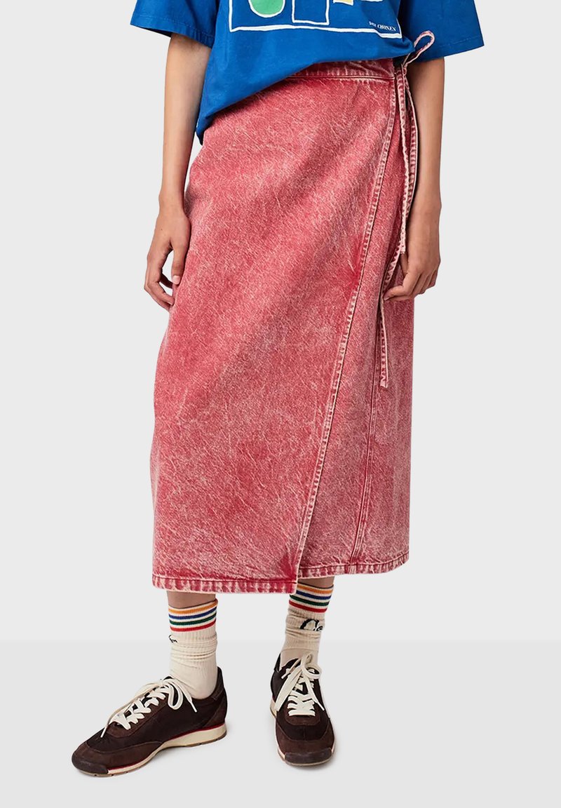Jupe en denim rouge enveloppante avec une texture décolorée, dotée d'une fermeture à nouer sur le côté et d'un ourlet droit. Assortie à des baskets sombres et des chaussettes rayées.