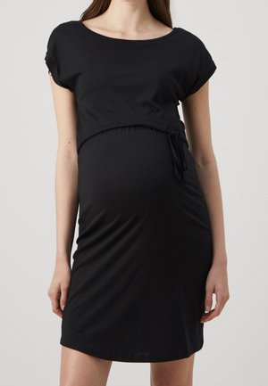 Kobieta w ciąży ubrana w dopasowaną czarną sukienkę z krótkim rękawem, z wciętą talią i detalem wiązania z boku, stoi na jednolitym tle.