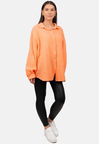 Camicia in cotone oversize, abbottonata, di un vivace arancione con una finitura testurizzata e colletto, abbinata a leggings neri e sneaker bianche.