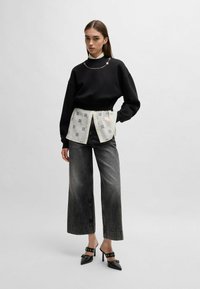 Zwarte oversized sweatshirt, gelaagd met een witte shirt met grafische prints, gecombineerd met wijde, vervaagde grijze jeans en puntige zwarte hakken.