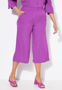Pantalones cortos de pierna ancha en color púrpura, confeccionados con una tela ligera y texturizada, que presentan una cinturilla elástica y bolsillos laterales, combinados con sandalias de tacón bloque a juego.