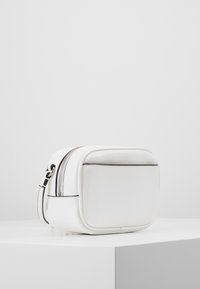 Compatta pochette in pelle bianca con tasca frontale e cerniera superiore, posata su un piedistallo bianco davanti a uno sfondo uniforme.