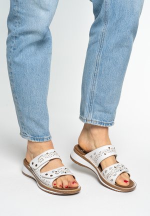 Füße mit weißen, perforierten Ledersandalen mit verstellbaren Riemen und hellblauen Jeans vor einfachem Hintergrund.