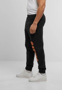 Black sweatpants feature an orange graphic logo on the left leg, elastic waistband, and cuffed ankles, paired with white sneakers.