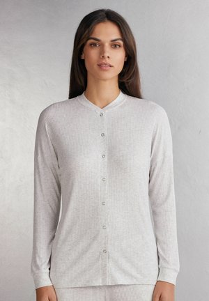 Vrouw met lang bruin haar die een lichtgrijs geribd overhemd met knopen en lange mouwen draagt, staat voor een effen achtergrond.