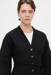Svart, knelang cardigan laget av tykk stoff med V-hals, lange ermer, knyting i midjen og store knapper foran.