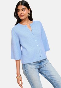 Jasnoniebieski sweter robiony na drutach z okrągłym dekoltem, półrękawami i zapięciem na guziki. Posiada subtelną teksturę i prążkowany dół.