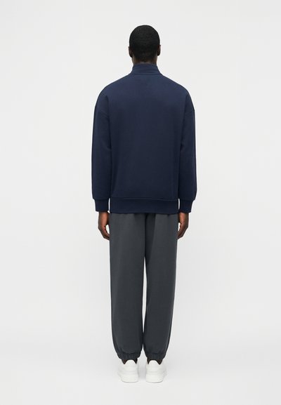 Homme debout, de dos, portant un sweat-shirt bleu marine, un pantalon de jogging gris foncé avec des poignets élastiques, et des baskets blanches, sur un fond uni.