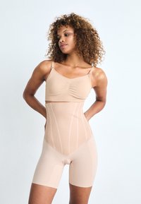 Le body sculptant beige présente une texture lisse, un design structuré, des bretelles ajustables et des détails de coutures verticales. Stylé de manière fonctionnelle avec une coupe short.