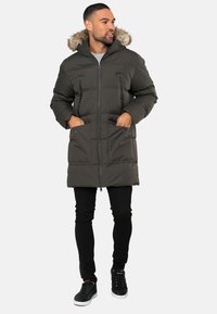 Olijfgroene gewatteerde parka met een bontgevoerde capuchon, ritssluiting aan de voorkant, twee ritszakken op de borst en twee grote broekzakken. Gelaagd weefsel.