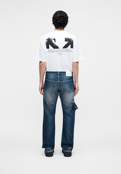 Homme portant un t-shirt blanc avec un motif de flèche noire et du texte au dos, accompagné d'un jean bleu ample et de chaussures noires, vu de dos.