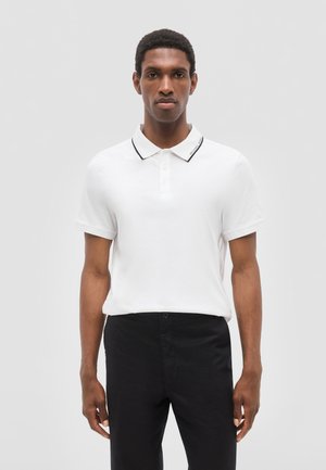 Hombre de pie mirando hacia adelante, vestido con una camisa polo blanca de manga corta con ribete negro en el cuello y pantalones negros.