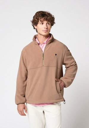 Jonge man met een lichtbruine halfrits fleece, handen in de zakken, roze shirt eronder, en witte broek, kijkt iets naar de zijkant.