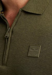 Olive zelena polo majica s patentnim zatvaračem na ovratniku, teksturirana tkanina i metalna oznaka s natpisom "PME LEGEND" na prsima.